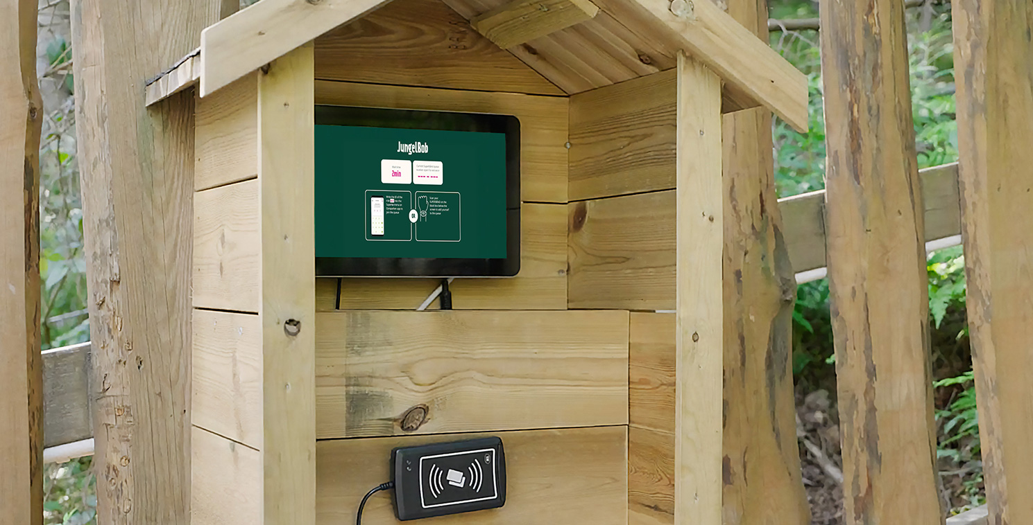 Ein Besucher interagiert mit einem wetterfesten Touchscreen-Kiosk in einem Holzgehäuse in einem Freizeitpark und demonstriert Self-Service-Technologie für den Ticketverkauf.